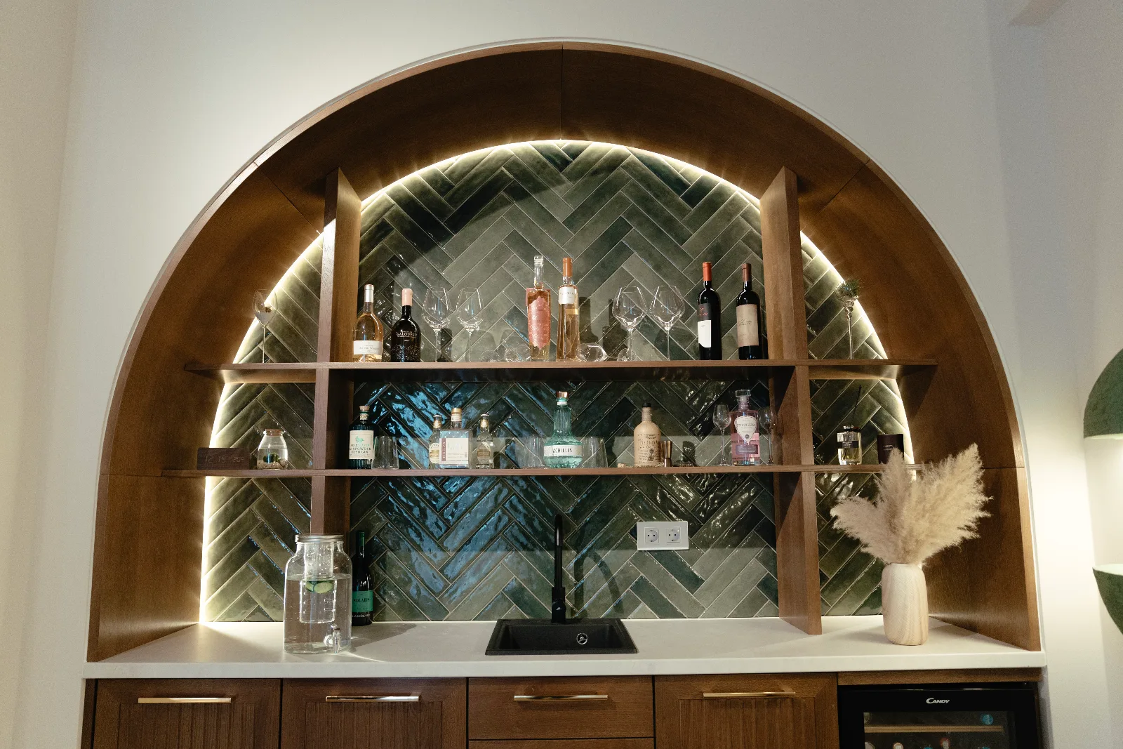 Arched oak bar niche with backlit shelving over green herringbone tiles